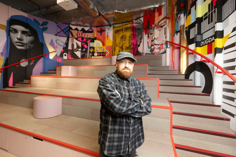 Artist Alec Saunders stands in front of his colourful wall mural of faces and other patterns at the Dugdale Arts Centre