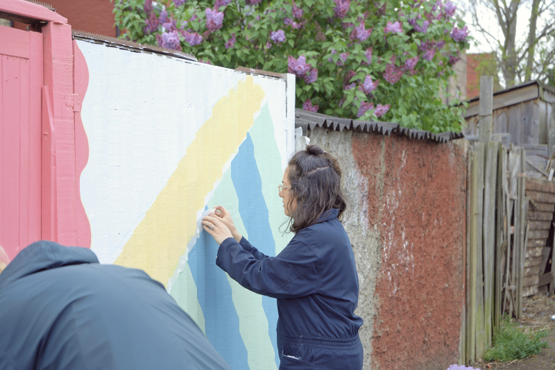 A woman applies a geometric design to a whitewashed wooden fence