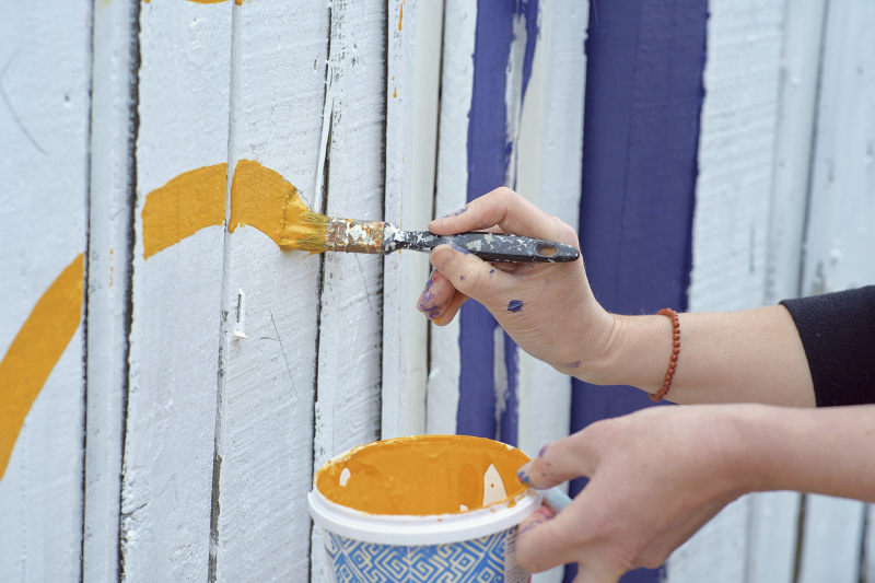 A close-up of hands painting a yellow colour onto a whitewashed wooden fence