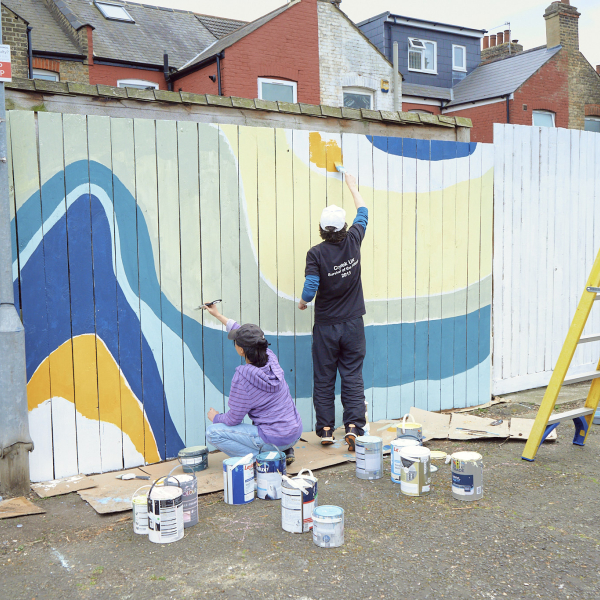 A man and woman paint a back fence with a colourful, wavy design