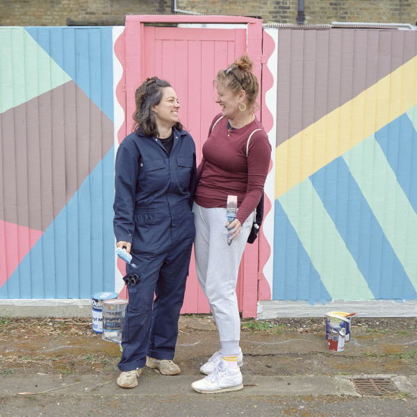 Two women smile at each other and hold paintbrushes in front of a colourfully painted garden fence