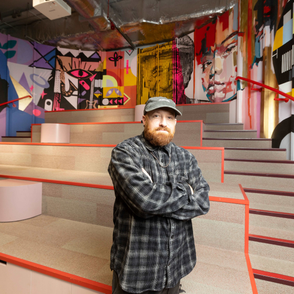 Alec Saunders stands in front a large, colourful wall mural on a curved back wall behind a stage at Dugdale Arts Centre