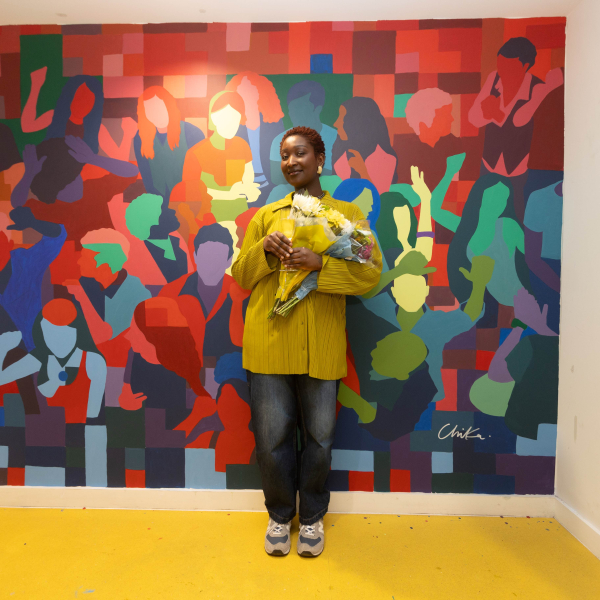 A woman stands in front of a colourful mural featuring coloured silhouettes of multiple figures and faces