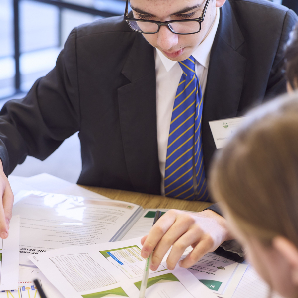 A student wearing glasses and a suit with a blue and yellow striped tie points to a document while working at a table with others.