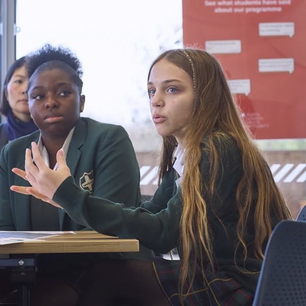A young person wearing a school uniform is speaking animatedly with their hands raised, while another person in uniform looks on intently