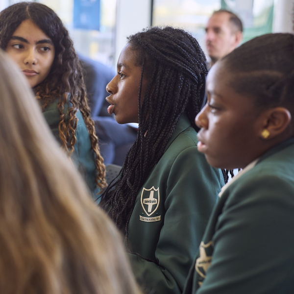 Three students in dark green blazers with a school crest are sitting and listening attentively. The student in the middle is speaking.