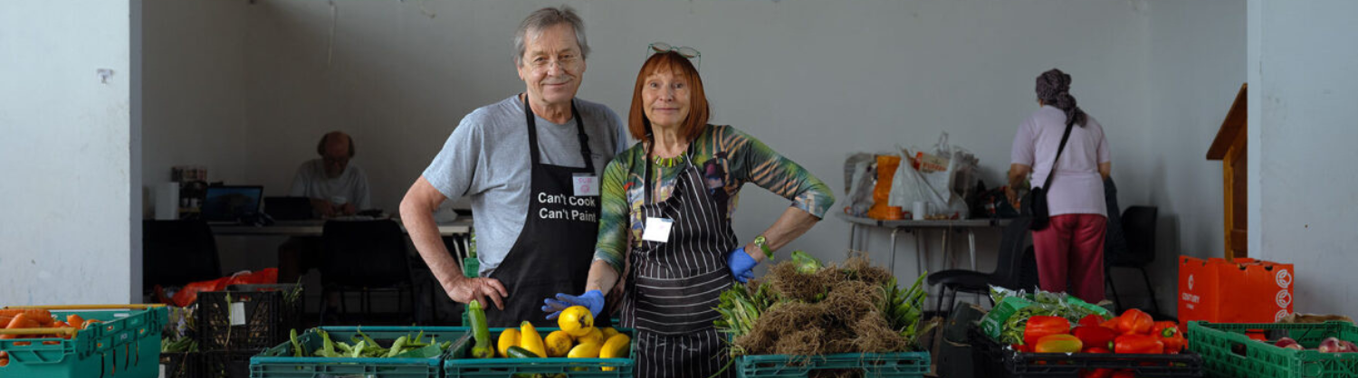 Two people stand behind cartons of fresh fruit and vegetables