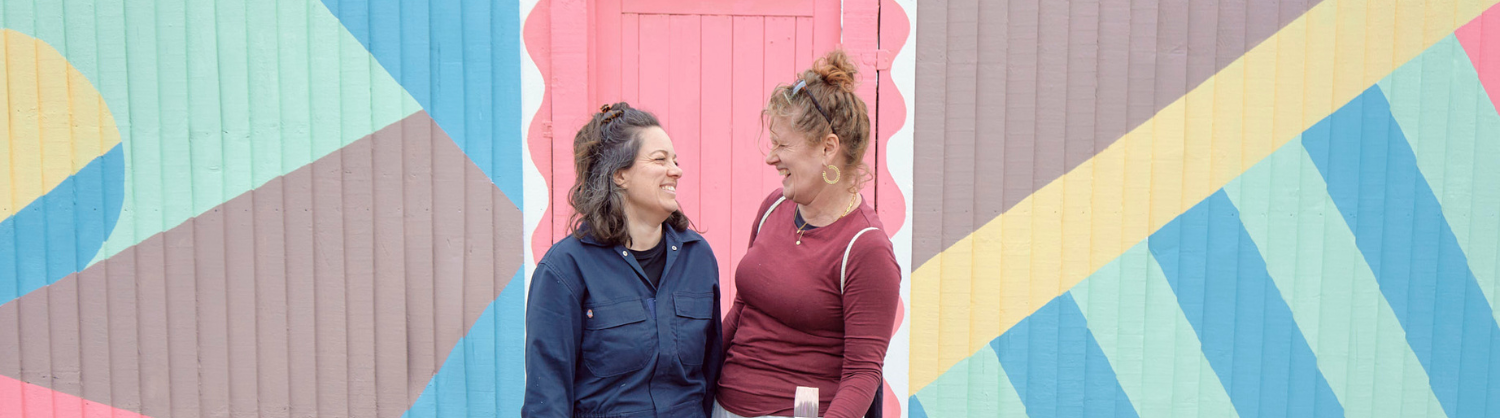 Two women smile at each other in front of a colourfully painted fence