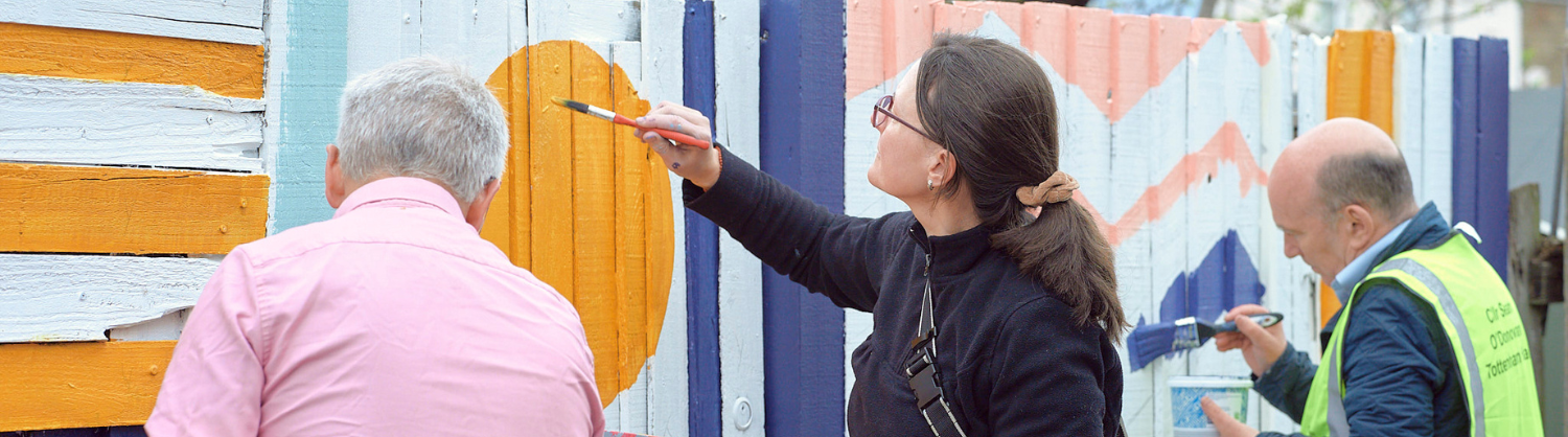 Three people paint a colourful geometric design onto a garden fence