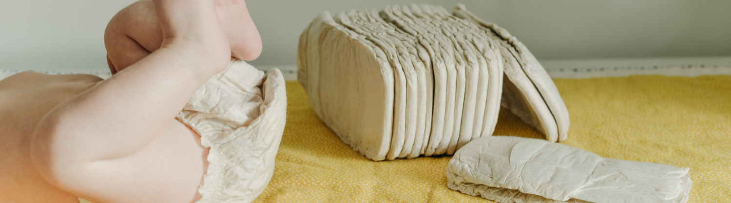 A baby in a nappy lies on a tabletop next to a stack of nappies