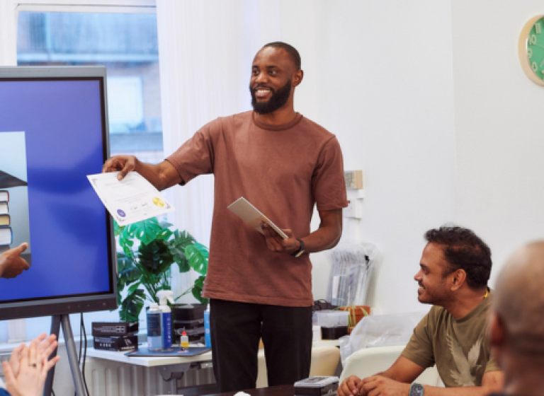 A man stands in front of a big TV screen and smiles, handing a certificate to an unseen person