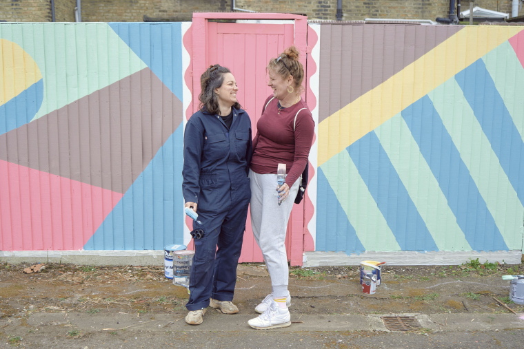Two women smile at each other in front of a colourful fence