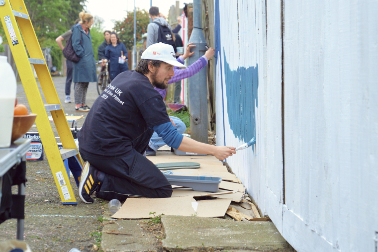 A man crouches down painting a blue pattern onto a wooden fence
