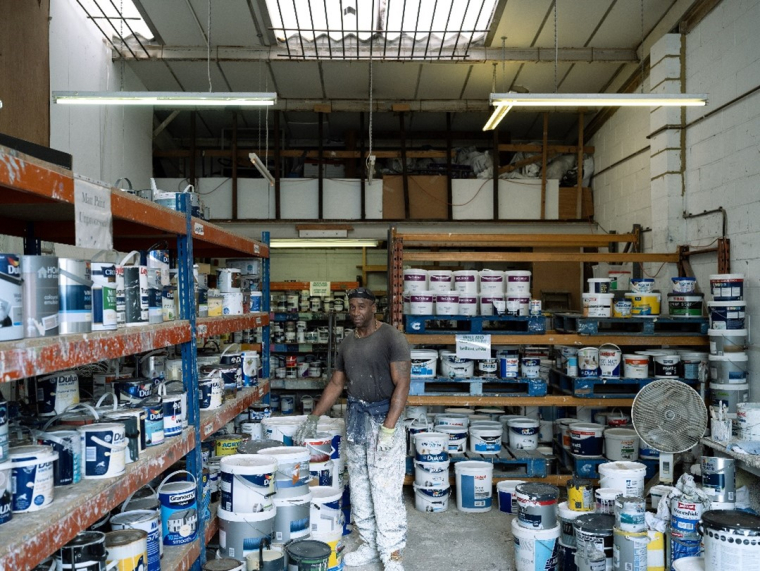A man stands in amongst numerous paint cans in a warehouse