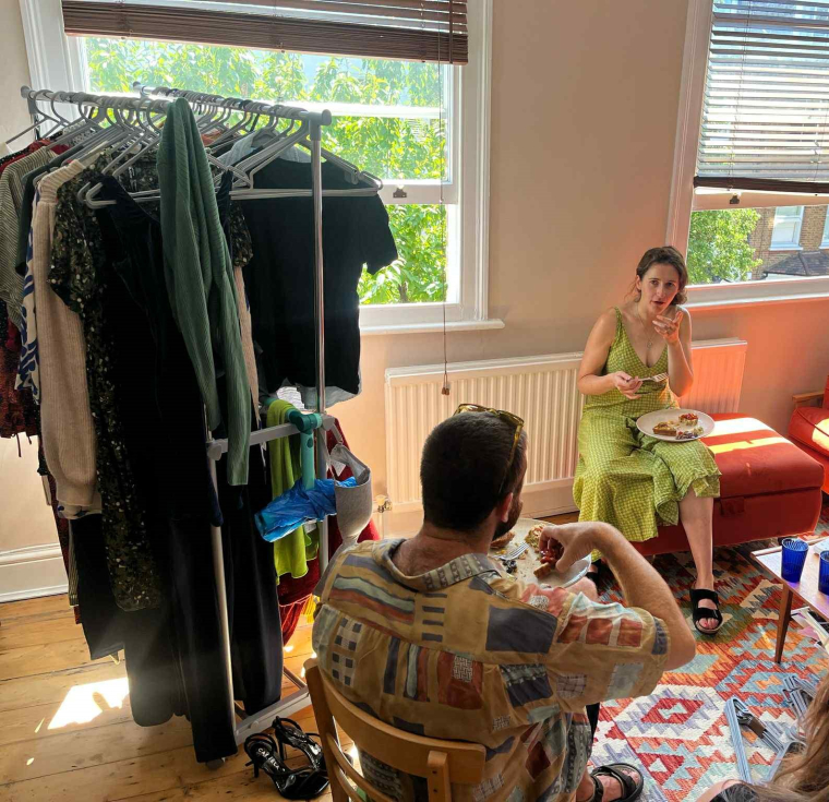 People eating and chatting next to a rail of clothes at a clothes swap