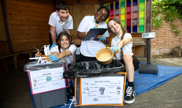 Four children sit behind their school bins, holding out items of rubbish and a clipboard, smiling at the camera