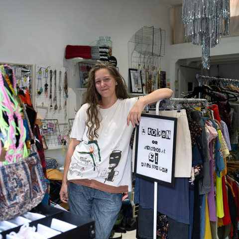 Image of a person standing inside clothing shop leaning against a clothing rail and smiling to the camera