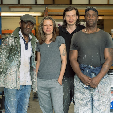 Image of four people standing inside shop surrounded by paint tins, smiling at the camera