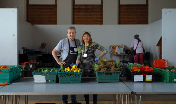 Two people stand behind cartons of fresh fruit and vegetables